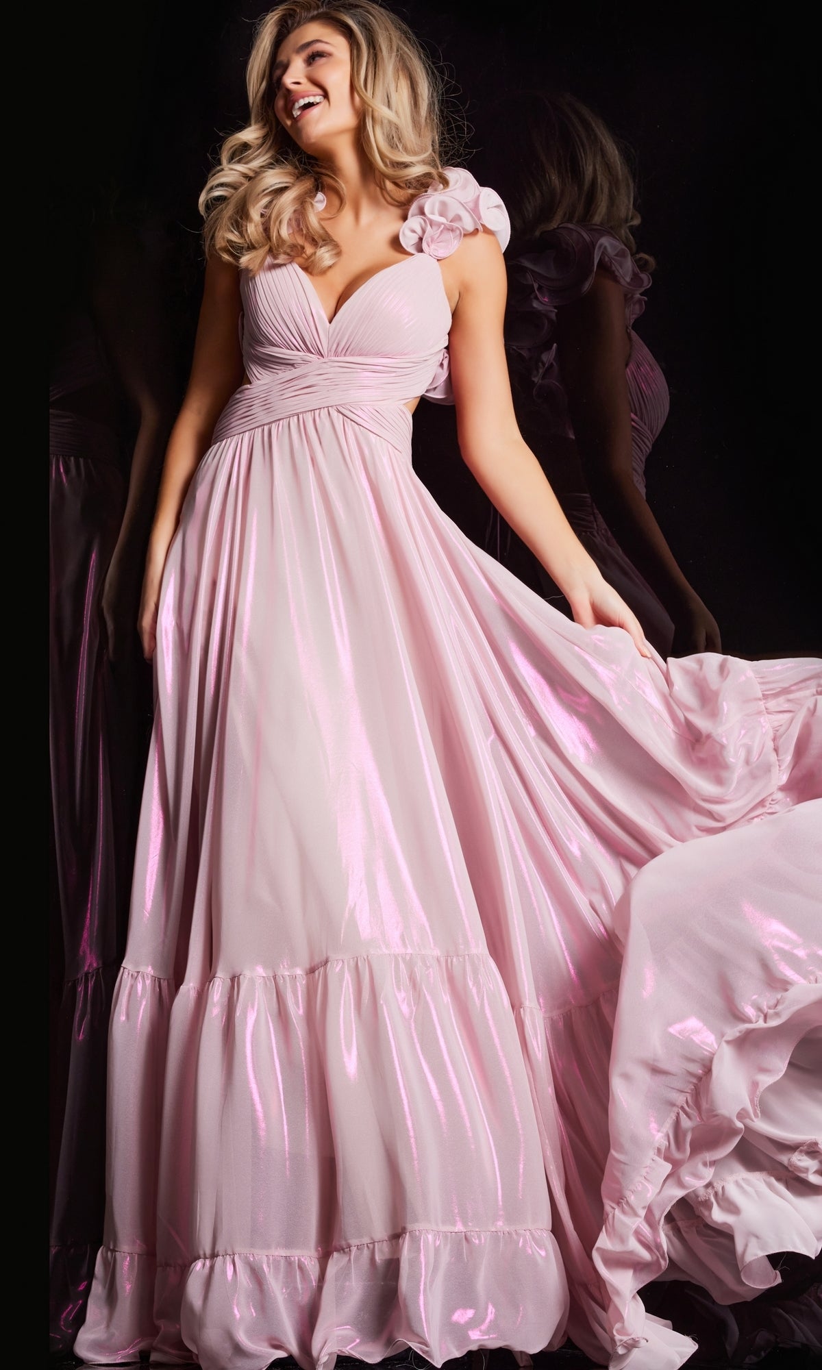 A woman wears the Jovani Ruffle-Strap Long Metallic Prom Dress 26248 in shiny light pink, featuring ruffled straps and a flowing A-line skirt, holding out one side of the dress against a dark background.